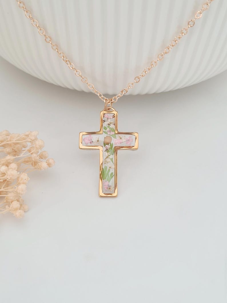 May include: A gold-colored cross pendant necklace. The cross is filled with small pink and white flowers and greenery. The necklace chain is also gold-colored. The pendant hangs against a white background.