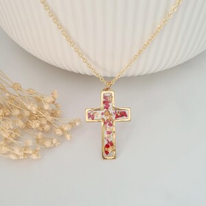 May include: A gold-colored necklace featuring a cross pendant. The cross is filled with red and white floral elements, encased in a gold frame. The chain is delicate and gold, creating a simple yet elegant design. The necklace is displayed on a white surface.