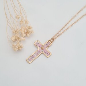 May include: A gold chain necklace with a delicate cross pendant. The cross is filled with pink pressed flowers.