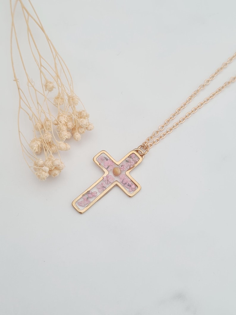 May include: A gold chain necklace with a delicate cross pendant. The cross is filled with pink pressed flowers.