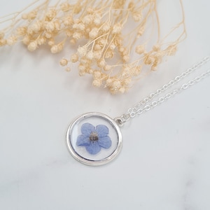 May include: A silver necklace with a round pendant featuring a pressed blue flower encased in clear resin.