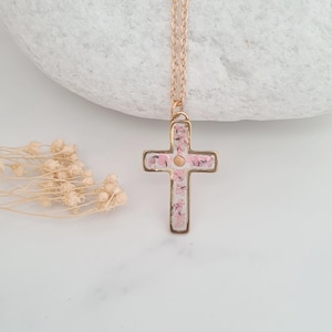 May include: A gold chain necklace with a delicate cross pendant. The cross is filled with pink pressed flowers.