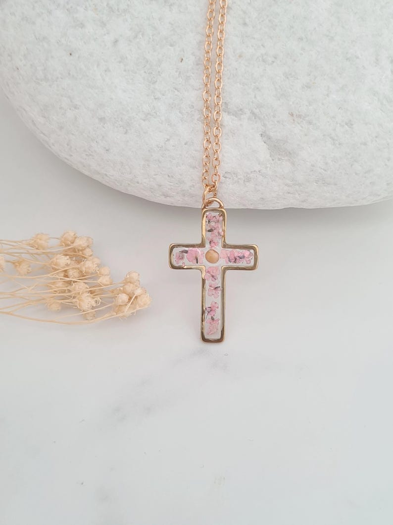 May include: A gold chain necklace with a delicate cross pendant. The cross is filled with pink pressed flowers.