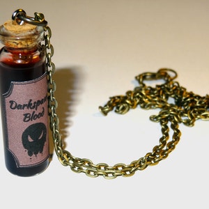 May include: A glass bottle necklace with a cork stopper and a brown label that reads "Darkspawn Blood". The label has a black skull graphic. The necklace is made of a gold chain.