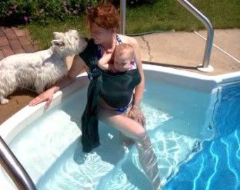 May include: A person in a pool with a baby in a dark green carrier. A white dog stands nearby. The pool has blue water and a metal handrail. The scene is outdoors on a sunny day.