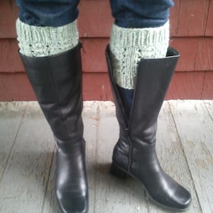 May include: A pair of black leather knee-high boots with a square toe. The boots are worn with light green knitted leg warmers.