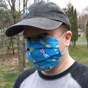 May include: A blue face mask with a dragonfly pattern. The mask is made of fabric and has white elastic ear loops. The person in the image is also wearing a gray baseball cap.