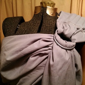 Può includere: Un'avvolgibile per neonati grigia con un anello argentato. L'avvolgibile è realizzata in un tessuto morbido e traspirante ed è progettata per essere comoda sia per il bambino che per chi la indossa.