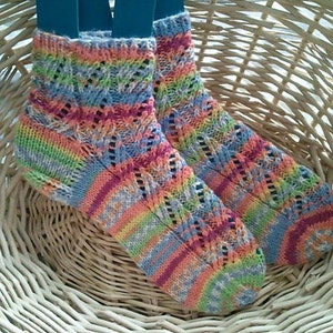 May include: A pair of colorful, hand-knitted socks with a rainbow pattern. The socks feature a diamond-shaped openwork design and are displayed in a woven basket. The color palette includes shades of pink, blue, green, and yellow.