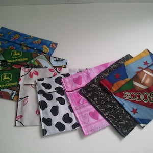 Puede incluir: Una colección de ocho bolsas de tela para aperitivos con varios diseños, incluyendo John Deere, fútbol y estampado de vaca. Las bolsas están todas dobladas y dispuestas sobre una superficie blanca.