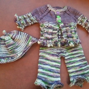 Puede incluir: Un conjunto de punto para bebé en tonos de morado, verde y blanco. El conjunto incluye un cárdigan con cierre de botón, pantalones y un gorro a juego.