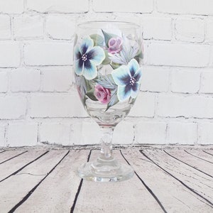 May include: A clear glass wine glass with a hand-painted design of blue and pink flowers. The flowers are painted in a realistic style and have green leaves.