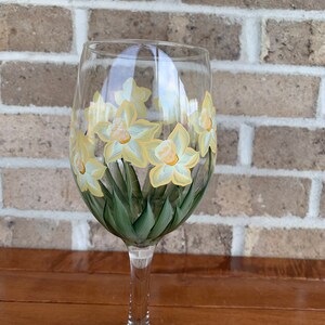 May include: A clear wine glass hand-painted with yellow and white daffodils and green leaves. The floral design wraps around the glass, creating a spring-like aesthetic. The glass sits on a wooden surface against a brick background.