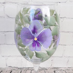 May include: A clear glass wine glass with a hand-painted purple pansy flower design. The flower has a yellow center and green leaves.