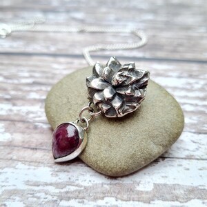 Peut inclure: Un collier en argent avec un pendentif en forme de fleur texturée et une pierre précieuse rose pendante. Le collier est présenté sur une pierre grise.