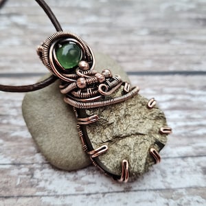 May include: A copper wire wrapped pendant featuring a green gemstone and a gold-toned pyrite stone. The pendant is attached to a brown leather cord.