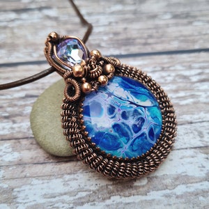 May include: A copper wire wrapped pendant featuring a blue and white marbled resin cabochon and a clear iridescent crystal. The pendant is suspended from a brown leather cord.