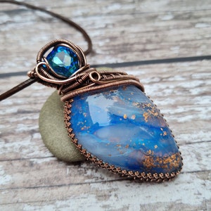 Può includere: Una collana con pendente realizzata a mano con una pietra blu e bianca con scaglie d'oro, racchiusa in filo di rame. Una gemma blu sfaccettata si trova sopra il pendente. La collana è appesa a un cordino di cuoio marrone, mostrando un design artistico unico.