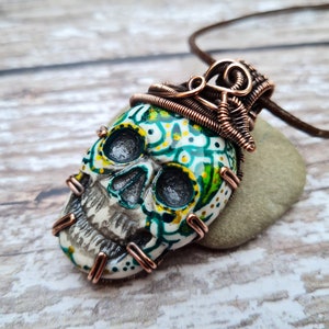 May include: A white skull pendant with green and yellow painted details, wrapped in copper wire, and hanging from a brown cord.