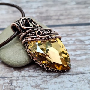 May include: A handcrafted pendant necklace with a large, faceted, amber-coloured crystal. The crystal is encased in intricate copper wire, suspended from a brown leather cord. The pendant rests on a small, grey stone.