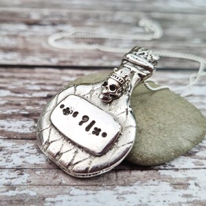 May include: Silver necklace with a pendant shaped like a bottle with a skull and crown on top. The bottle has a label that says "?" with a dot on either side.