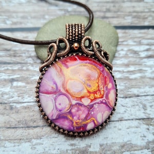 May include: A copper wire wrapped pendant necklace with a round, pink, red and gold abstract resin cabochon.