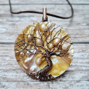 Peut inclure: Un pendentif en fil de cuivre en forme d'arbre de vie sur fond de nacre. Le pendentif présente un design filaire détaillé et de petits accents de perles.
