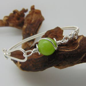 Peut inclure: Un bracelet jonc argenté avec une perle ronde vert vif au centre, entourée de fil d'argent. Le bracelet est doté d'une fermeture à crochet et de volutes décoratives. Il est présenté sur un morceau de bois flotté.