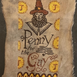 May include: A rectangular piece of grey linen fabric featuring a cross-stitch design of a person in a brown hat, surrounded by yellow circular coins marked with the letter J. The text reads Penny for the Guy, with decorative borders.