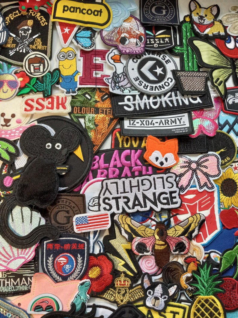 May include: A collection of colourful iron-on patches featuring various designs, including cartoon characters, animals, logos, and text. The patches are arranged in a random pattern on a white background.