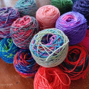 May include: A collection of colorful yarn balls in various shades of blue, green, pink, red, purple, and black. The yarn is arranged in a circular pattern on a wooden surface.