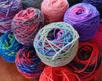 May include: A collection of colorful yarn balls in various shades of blue, green, pink, red, purple, and black. The yarn is arranged in a circular pattern on a wooden surface.