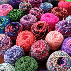 May include: A collection of colorful yarn balls in various shades of pink, red, green, blue, purple, and black. The yarn balls are arranged in a random pattern on a wooden surface.