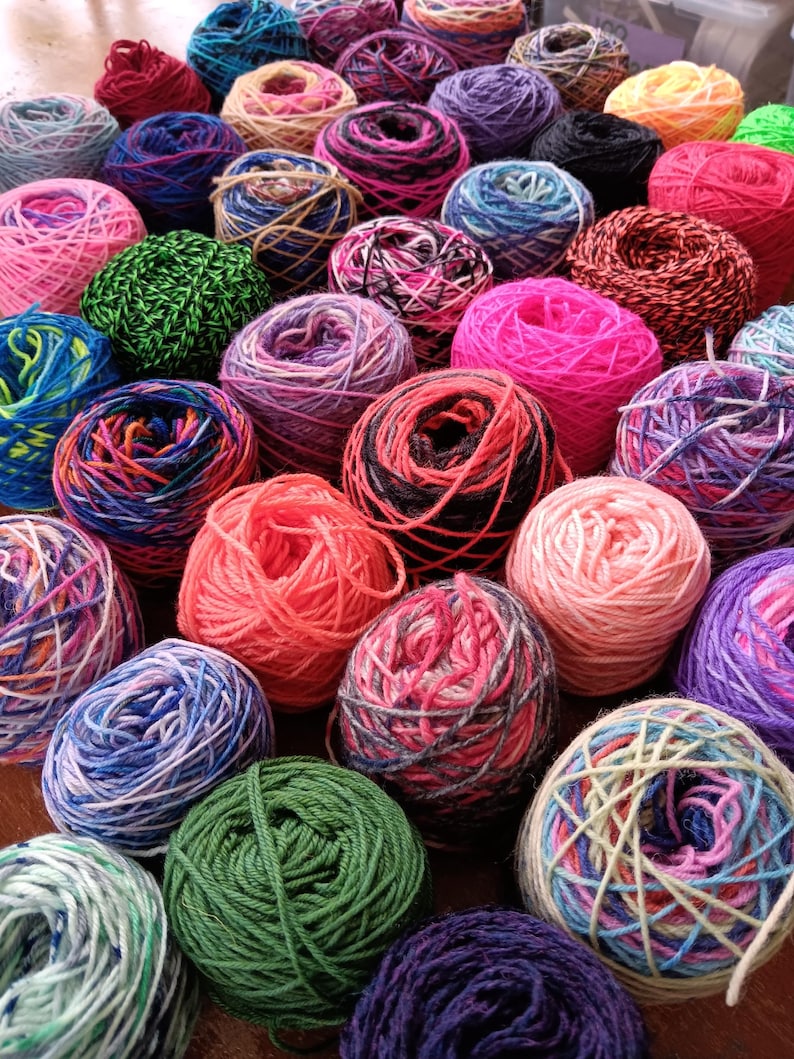 May include: A collection of colorful yarn balls in various shades of pink, red, green, blue, purple, and black. The yarn balls are arranged in a random pattern on a wooden surface.