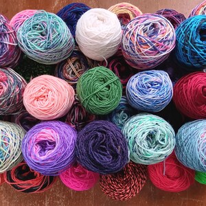 May include: A collection of colorful yarn balls in various shades of blue, green, pink, red, and white. The yarn is arranged in a circular pattern on a wooden surface.