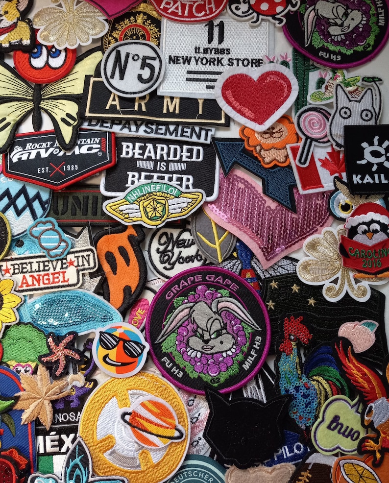 May include: A collection of colourful embroidered iron-on patches featuring various designs, including a butterfly, a heart, a rooster, a cartoon bunny, a penguin, a star, a planet, and a sunflower. The patches are made of fabric and have a variety of colours and textures.
