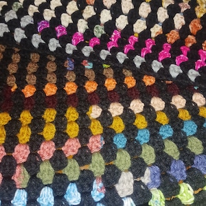 May include: Close-up of a handmade crochet blanket. The blanket features a granny square design with a black background and colorful squares in shades of pink, orange, green, blue, and yellow. The texture is visible.