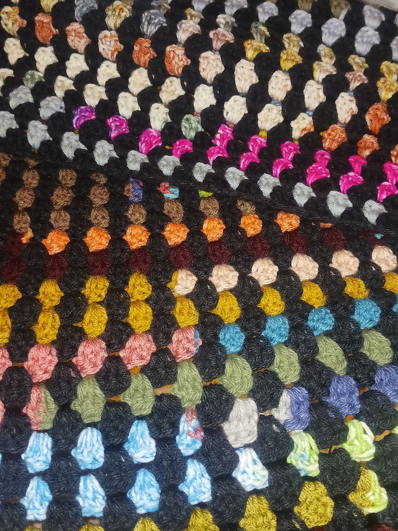 May include: Close-up of a handmade crochet blanket. The blanket features a granny square design with a black background and colorful squares in shades of pink, orange, green, blue, and yellow. The texture is visible.