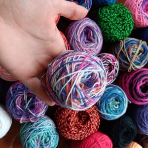 May include: A close-up of a variety of colorful yarn balls, including purple, blue, green, red, white, and orange. The yarn is arranged in a random pattern, and some of the balls are partially obscured by a hand.
