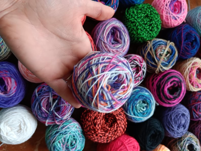 May include: A close-up of a variety of colorful yarn balls, including purple, blue, green, red, white, and orange. The yarn is arranged in a random pattern, and some of the balls are partially obscured by a hand.