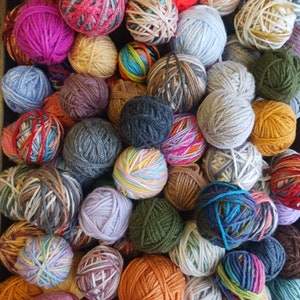 May include: A close-up of numerous balls of yarn in a range of colours, including pink, orange, blue, and green. The yarn balls are tightly wound and packed together, displaying a variety of textures and colour combinations, suitable for knitting or crochet.