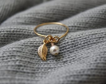 May include: A gold ring featuring a leaf charm and a white pearl charm. The ring is set against a grey, textured background. The leaf charm has detailed veining.