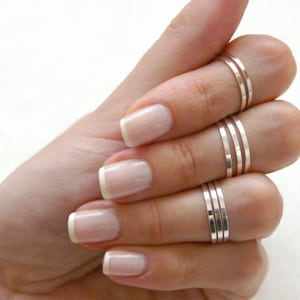 May include: A hand wearing multiple silver stacking rings on the ring finger. The nails are painted with a light pink polish and have a white tip.