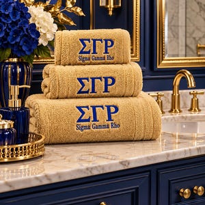 May include: A stack of three tan towels featuring blue embroidered Greek letters Sigma Gamma Rho. The top and bottom towels include the full text Sigma Gamma Rho in blue embroidery. The towels are folded neatly and display a textured fabric surface.