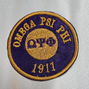 May include: Embroidered patch with the words "OMEGA PSI PHI" in gold lettering on a purple background. The center features the Greek letters "ΩΨΦ" in gold, with the year "1911" below. The patch has a gold border.