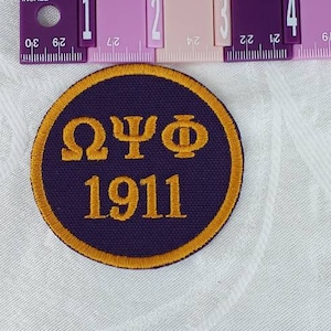 May include: A purple and gold embroidered patch with the letters "ΩΨΦ" and the year "1911" in the center.