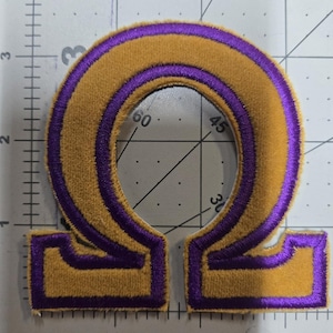 May include: An embroidered Greek letter Omega patch featuring a gold-colored center with a purple border. The patch is positioned on a cutting mat with visible inch markings, showing the letter measures approximately 3 inches in height.