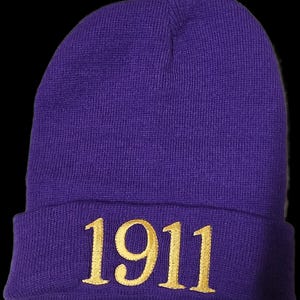 May include: A purple knit beanie with the gold number "1911" embroidered on the folded brim. The hat is made of a soft, textured material and is set against a black background.