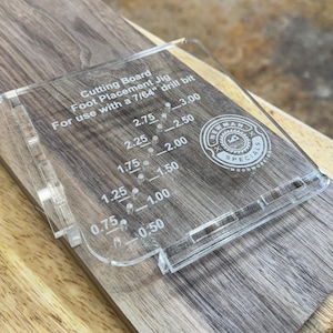 May include: A clear acrylic cutting board foot placement jig with white printed text reading Cutting Board Foot Placement Jig For use with a 7/64 drill bit and numerical measurements from 0.50 to 3.00 inches, featuring a circular Newman Specials logo.