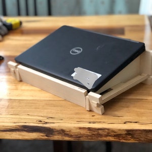 May include: A wooden laptop stand with a black laptop resting on top. The stand is made of light wood and has a simple design. The laptop has a Dell logo on the lid.